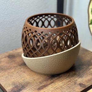 May include: A brown, openwork decorative planter with a geometric design, set atop a beige, textured base. The planter sits on a wooden surface, showcasing its intricate pattern and color contrast.