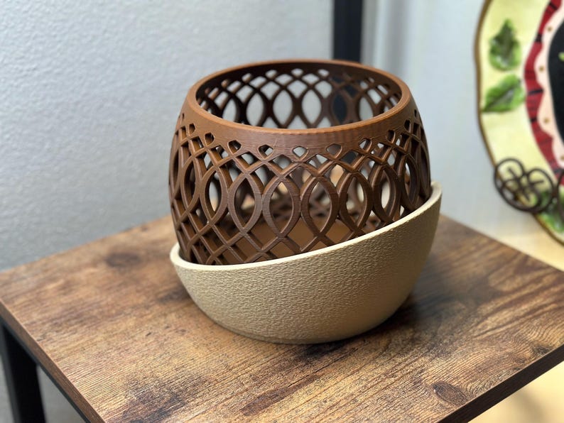 May include: A brown, openwork decorative planter with a geometric design, set atop a beige, textured base. The planter sits on a wooden surface, showcasing its intricate pattern and color contrast.