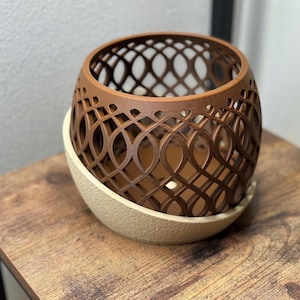 May include: A brown, openwork decorative planter with an intricate geometric design. The planter sits on a beige, oval-shaped base. The item is displayed on a wooden table.