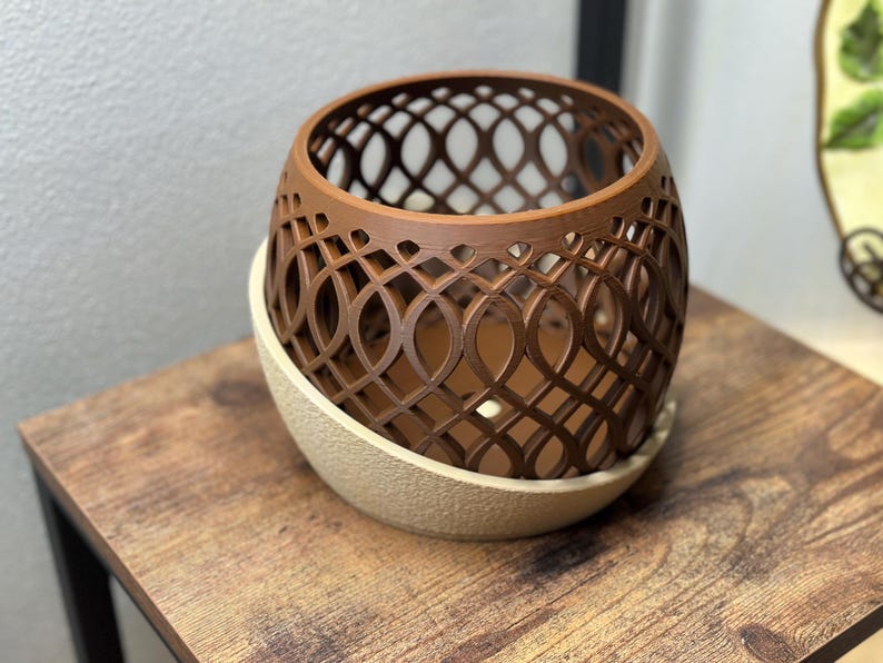 May include: A brown, openwork decorative planter with an intricate geometric design. The planter sits on a beige, oval-shaped base. The item is displayed on a wooden table.