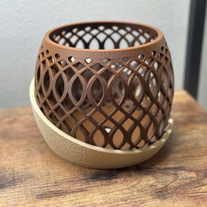 May include: A brown, openwork decorative planter with an intricate geometric design. The planter sits on a beige, rounded base. The design is a repeating pattern of intersecting ovals and lines, creating a lattice effect. The planter is made of a solid material.