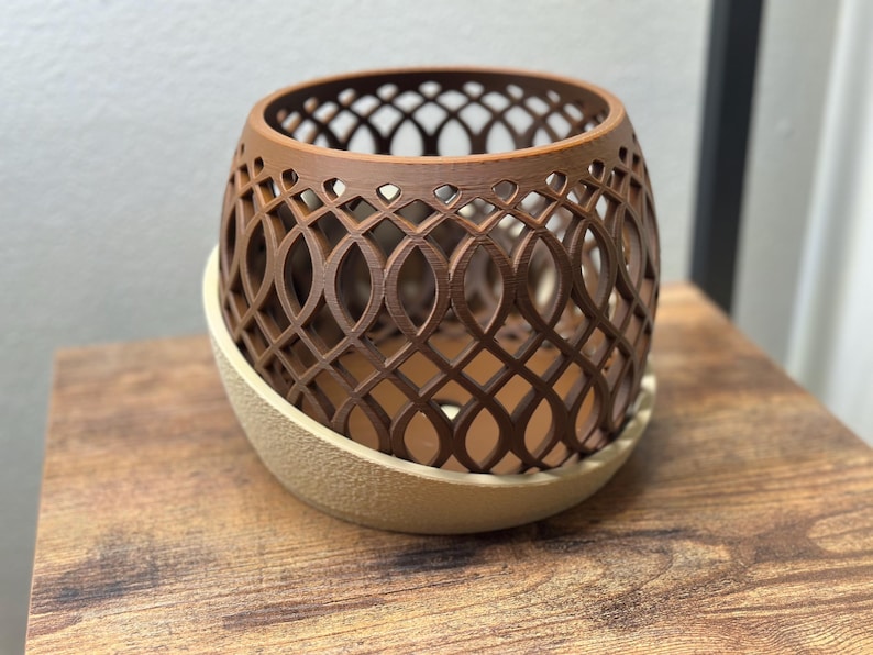 May include: A brown, openwork decorative planter with an intricate geometric design. The planter sits on a beige, rounded base. The design is a repeating pattern of intersecting ovals and lines, creating a lattice effect. The planter is made of a solid material.