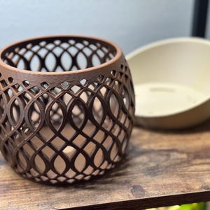 May include: A brown, openwork, spherical decorative object with a repeating oval pattern. Beside it is a cream-colored, oval-shaped bowl. Both items are on a wooden surface.