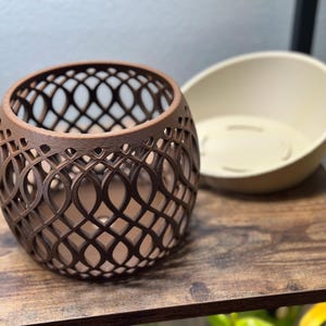 May include: A brown, openwork, spherical decorative container with an intricate lattice design. Beside it is a cream-colored, oval-shaped planter. Both items sit on a dark wooden shelf, suggesting a home decor theme.