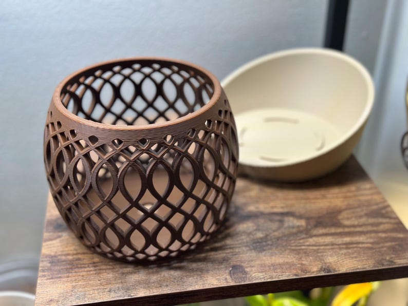 May include: A brown, openwork, spherical decorative container with an intricate lattice design. Beside it is a cream-colored, oval-shaped planter. Both items sit on a dark wooden shelf, suggesting a home decor theme.