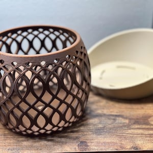 May include: A brown, openwork, decorative vase with an intricate pattern, next to a cream-colored, oval-shaped bowl. The vase has a rounded shape and is made of a dark brown material. The bowl is smooth and has a simple design.