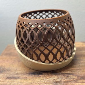 May include: A brown, openwork planter with an intricate, repeating pattern. The planter sits on a beige, rounded base. The design is a decorative element for home decor, suitable for holding small plants or other items.