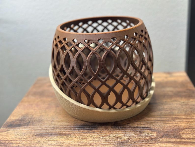 May include: A brown, openwork planter with an intricate, repeating pattern. The planter sits on a beige, rounded base. The design is a decorative element for home decor, suitable for holding small plants or other items.