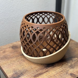 May include: A brown, openwork decorative planter with a lattice design, sitting on a beige, textured base. The planter is on a wooden table. The design is a series of intersecting ovals, creating an intricate pattern.