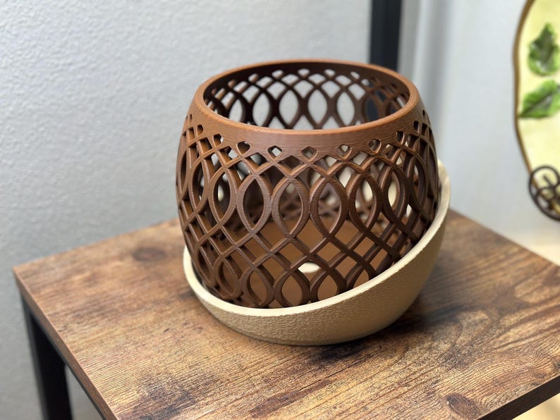 May include: A brown, openwork decorative planter with a lattice design, sitting on a beige, textured base. The planter is on a wooden table. The design is a series of intersecting ovals, creating an intricate pattern.