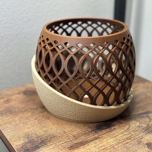May include: A brown, openwork decorative planter with a geometric design. The planter sits on a beige, textured saucer. The planter is on a wooden table.