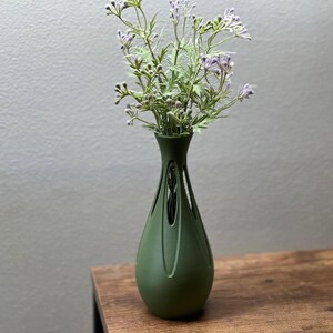 A green vase with purple flowers sits on a wooden surface against a plain gray wall.