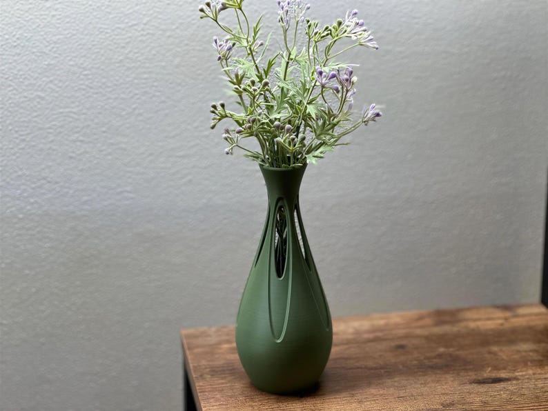 A green vase with purple flowers sits on a wooden surface against a plain gray wall.