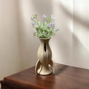 May include: A beige ceramic vase with a rocket ship design, holding a bouquet of lavender-colored flowers. The vase has a unique shape with fin-like structures on the sides. The flowers have green stems and small purple blooms.
