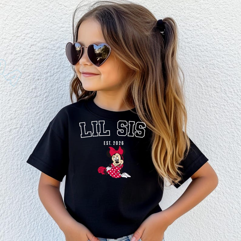 May include: A black t-shirt featuring the text "LIL SIS EST. 2026" in white. The shirt also has a cartoon Minnie Mouse graphic in a red polka dot dress and bow. The girl is wearing heart-shaped sunglasses.