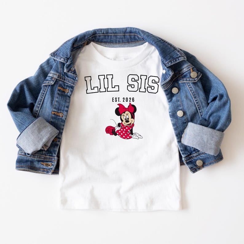 May include: A white t-shirt with the text "LIL SIS EST. 2026" in black. The shirt features a graphic of Minnie Mouse in a red polka dot dress and bow. A blue denim jacket is draped over the shirt.