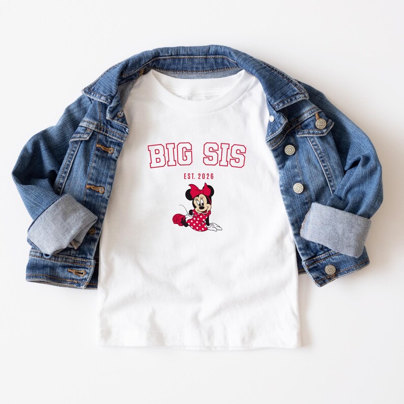 Pu&ograve; includere: Una t-shirt bianca a maniche corte con la scritta rossa BIG SIS ed EST. 2026 sopra un'immagine di Minnie Mouse con un vestito a pois rossi e un fiocco, indossata sotto una giacca di jeans blu con bottoni in metallo e maniche lunghe risvoltate.