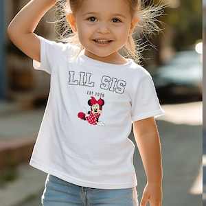 May include: White t-shirt with the text "LIL SIS" in black block letters and "EST. 2026" below. The shirt features a Minnie Mouse graphic in red, black, and white. The child is wearing blue jeans.