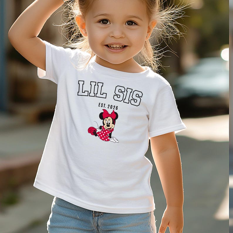 May include: White t-shirt with the text "LIL SIS" in black block letters and "EST. 2026" below. The shirt features a Minnie Mouse graphic in red, black, and white. The child is wearing blue jeans.