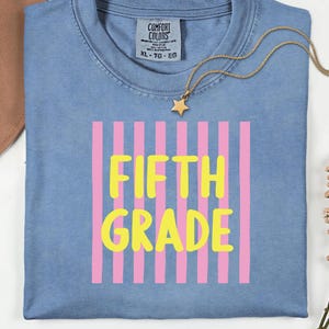 May include: A blue crew neck t-shirt featuring a graphic of vertical pink stripes with the yellow text FIFTH GRADE centered on the front. A gold-toned chain necklace with a small star pendant rests on the neckline of the shirt.