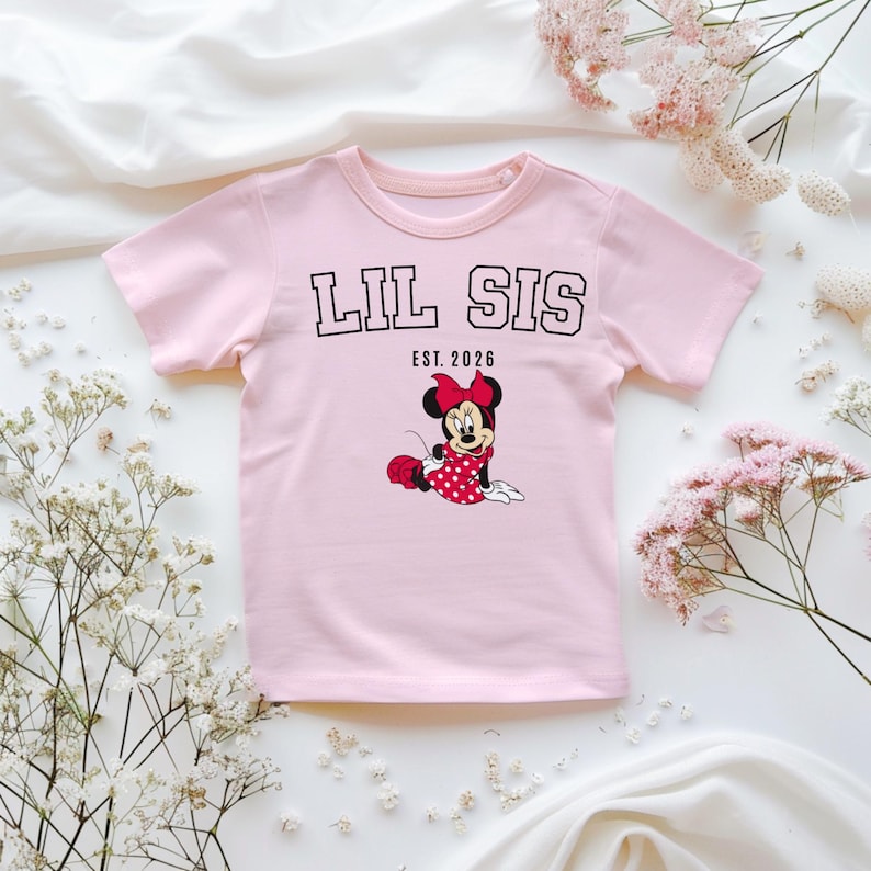 May include: A pale pink t-shirt featuring the text "LIL SIS EST. 2026" in black. Below the text is a cartoon Minnie Mouse in a red polka dot dress and bow. The shirt is displayed on a white surface with floral decorations.