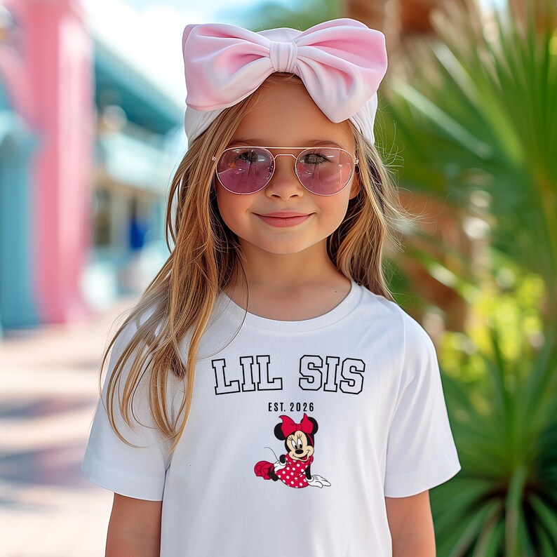 May include: A young girl wearing a white t-shirt with "LIL SIS EST. 2026" printed on it, along with a Minnie Mouse graphic. She is also wearing pink sunglasses and a large pink bow headband. The background is blurred.