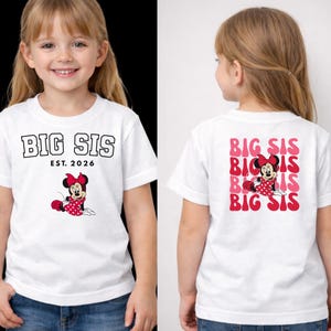 Pu&ograve; includere: Una t-shirt bianca a maniche corte con la scritta BIG SIS EST. 2026 e un'immagine di Minnie sul davanti. Il retro della maglietta presenta un motivo ripetuto della scritta BIG SIS in rosa e rosso con Minnie Mouse posizionata al centro.