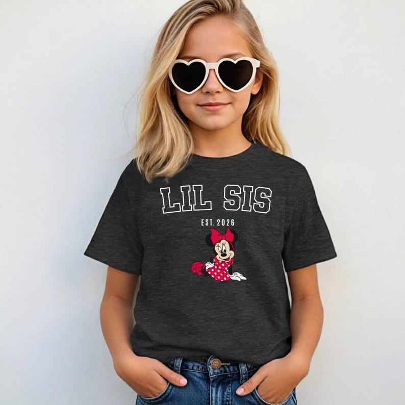 May include: A dark grey t-shirt with the text "LIL SIS EST. 2026" in white block letters. A cartoon Minnie Mouse in a red polka dot dress and bow is featured below the text. The girl is wearing heart-shaped sunglasses.