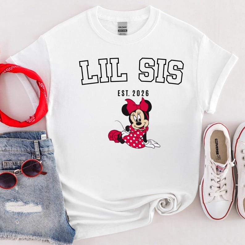 May include: White t-shirt with the text "LIL SIS EST. 2026" in black block letters. A cartoon Minnie Mouse in a red polka dot dress and bow is featured below the text. The shirt is paired with denim shorts, sunglasses, a red bandana, and white trainers.
