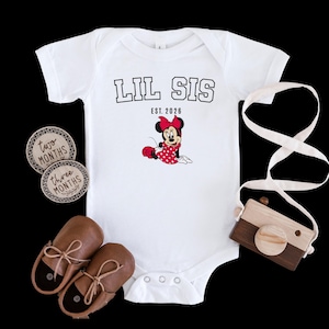 May include: White baby bodysuit with the text "LIL SIS" and "EST. 2026" above a Minnie Mouse graphic. The image also includes brown leather baby shoes, wooden monthly milestone markers, and a wooden toy camera.