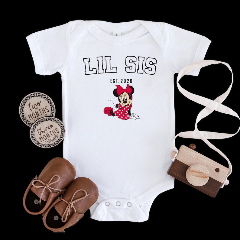 May include: White baby bodysuit with the text "LIL SIS" and "EST. 2026" above a Minnie Mouse graphic. The image also includes brown leather baby shoes, wooden monthly milestone markers, and a wooden toy camera.