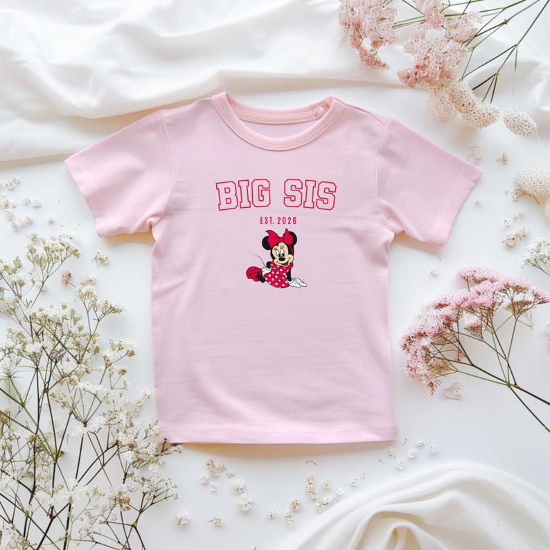 Pu&ograve; includere: Una t-shirt a maniche corte color rosa chiaro con la scritta rossa BIG SIS ed EST. 2026 sopra un'immagine di Minnie Mouse che indossa un vestito rosso a pois e un fiocco rosso. La maglietta ha un girocollo ed &egrave; in tessuto rosa chiaro.