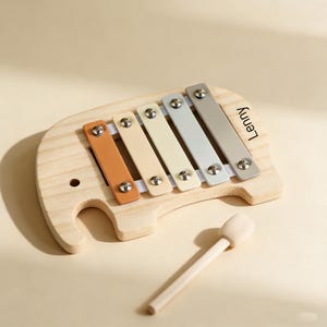 Könnte beinhalten: Holz-Xylophon in Elefantenform mit fünf farbigen Tasten: Orange, Creme, Hellgelb, Hellblau und Grau. Der Name "Lenny" ist auf der Seite aufgedruckt. Ein Holzschlägel ist enthalten.