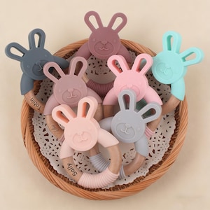 May include: A collection of colorful baby teethers in a woven basket. Each teether features a bunny-shaped silicone head in shades of pink, blue, gray, and purple, attached to a wooden ring. Some teethers have names printed on them.