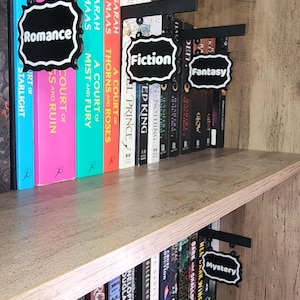 Könnte beinhalten: Holzregal mit schwarzen Metall-Regalmarkern mit den Genres "Romance", "Fiction", "Fantasy" und "Mystery". Die Bücher haben farbige Buchrücken und sind auf Holzregalen angeordnet. Der Text auf den Markern ist weiß.