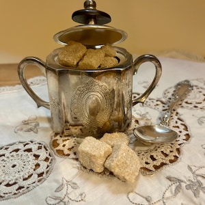 以下が含まれることがあります： 蓋付きの銀色の砂糖入れ。茶色の角砂糖が入っています。砂糖入れには2つの取っ手と装飾的なディテールがあります。小さな銀のスプーンが、刺繍された白い布の上に砂糖入れの隣に置かれています。