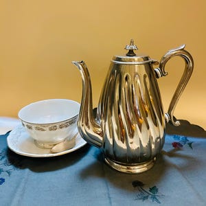 May include: A silver tea set with a fluted design, including a teapot, teacup, saucer, and spoon. The teacup and saucer are white with a gold floral pattern. The set is on a blue floral patterned tray.