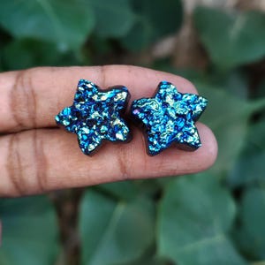 以下が含まれることがあります： 鮮やかな青、紫、緑の色合いを持つ、質感のある虹色のドゥルージー表面が特徴の星型スタッドピアス2点。クリスタルは光を反射するメタリックな仕上げで、光を捉えて多色に見える外観を作り出しています。