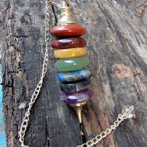May include: A pendulum with a silver chain and a stack of seven colorful gemstones. The gemstones are arranged in a column, with colors ranging from red to purple. The pendulum is designed for spiritual practices.