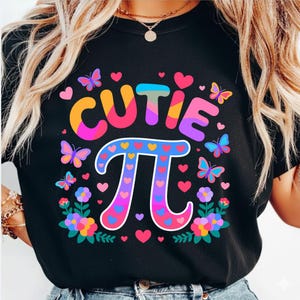 May include: Black t-shirt with a colorful graphic design. The design features the word "CUTIE" in rainbow colors above the mathematical symbol Pi, decorated with hearts, butterflies, and flowers. The shirt is a crew neck.
