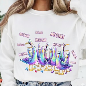Può includere: Felpa bianca con un disegno di un gruppo di uccelli dei cartoni animati colorati, ognuno con la scritta "MOM!" sopra. Gli uccelli hanno piume arcobaleno e sono su una superficie con piccole gemme.