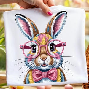 May include: White t-shirt featuring a colorful illustration of a bunny wearing pink glasses and a bow tie. The bunny has blue, yellow, pink, and brown fur. The design is centered on the shirt.