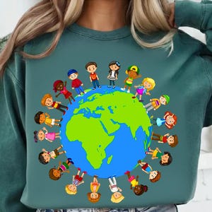 以下が含まれることがあります： 青と緑の地球を囲む子供たちのカラフルなグラフィックが特徴のティールスウェットシャツ。漫画の子供たちは多様で、さまざまな衣装を着ています。デザインはスウェットシャツの前面の中央に配置されています。