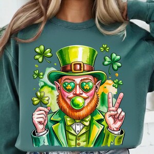 Può includere: Felpa verde con un design di un leprecauno dei cartoni animati. Il leprecauno ha una barba rossa, un cappello verde e occhiali da sole a forma di cuore con trifogli. Tiene in mano un trifoglio e fa il segno della pace. Il design include trifogli e una bolla verde.