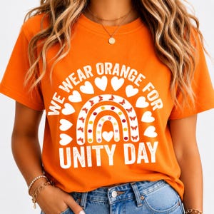 Puede incluir: Una camiseta naranja de manga corta con un estampado gráfico blanco de un arcoíris, corazones y mariposas. El texto "WE WEAR ORANGE FOR UNITY DAY" aparece en una fuente blanca, negrita y desgastada en el centro de la prenda.