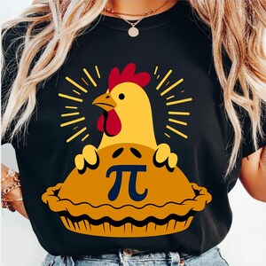 May include: Black t-shirt featuring a yellow chicken emerging from a pie with the mathematical symbol pi. The chicken has a red comb and wattle. The pie is golden brown with a crust. The design is centered on the shirt.