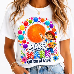 May include: A white t-shirt featuring a colorful graphic of a child holding a paintbrush and palette. The design includes a large orange circle, multi-colored dots, and the text "MAKE YOUR MARK ONE DOT AT A TIME!" in bold, vibrant lettering.