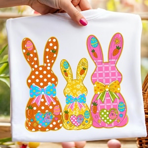 Op de afbeelding: Wit t-shirt met drie kleurrijke paashaasontwerpen. De konijnen hebben stippen, bloempatronen en een ruitpatroon. Elk konijn heeft een strik, worteldetails en paaseieren. Het t-shirt wordt door een hand vastgehouden.