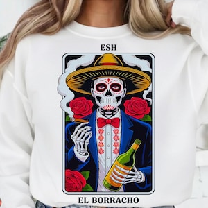 May include: White sweatshirt featuring a framed graphic of a skeleton in a sombrero, holding a bottle and cigar. The skeleton wears a blue suit and red bow tie, surrounded by red roses. Text reads "ESH" and "EL BORRACHO".