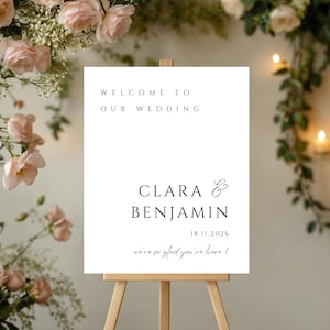 Puede incluir: Un letrero blanco de bienvenida de boda sobre un caballete de madera. El texto dice "Welcome to Our Wedding", con los nombres "Clara & Benjamin" y la fecha "18.11.2026". La frase "we're so glad you're here!" también está presente.